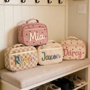 Puede incluir: Una colección de bolsas de almuerzo personalizadas en varios colores y patrones. Las bolsas presentan nombres como "Mia", "Jaxon", "Darcy" y "Ryder" en letras decorativas. Algunas bolsas tienen diseños de fresas o cuadros.