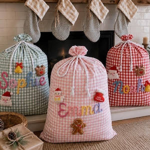 May include: Three fabric drawstring gift sacks in green, pink, and red gingham patterns. Each sack features embroidered names: Sophie, Emma, and Noah, decorated with plush holiday appliques like Santa, reindeer, gingerbread men, and bells.