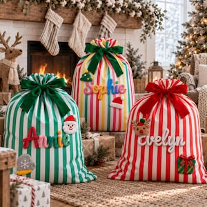 Puede incluir: Tres grandes sacos de regalo de tela con rayas verticales en verde, rojo y multicolor. Cada saco presenta un gran lazo de terciopelo en la parte superior y un nombre personalizado en relieve, acompañado de apliques de fieltro festivos como Santa, un árbol y un reno.