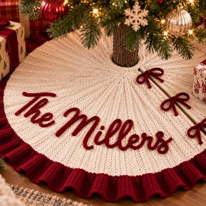 Op de afbeelding: Een ronde, crèmekleurige gebreide kerstboomrok met een donkerrode geplooide rand en drie bijpassende rode linten. De tekst "The Millers" is in donkerrood cursief schrift in het midden van de stof geborduurd.