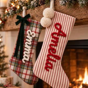 Puede incluir: Dos calcetines de Navidad colgados de una repisa de madera. Uno tiene un patrón de cuadros rojos y verdes con un lazo de terciopelo verde oscuro y el nombre Emma en blanco. El otro tiene rayas verticales rojas, dos pompones blancos y el nombre Amelia en rojo.