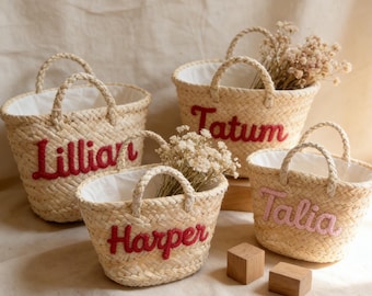 May include: Four woven straw baskets with handles, each featuring a different name embroidered in red or pink: Lillian, Tatum, Harper, and Talia. The baskets have white linings and are filled with dried flowers.