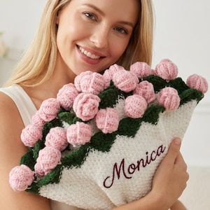 May include: A handmade bouquet of pink crochet flowers with green leaves and a white knitted wrap. The name "Monica" is embroidered in burgundy on the wrap. The bouquet is a unique, handcrafted gift.