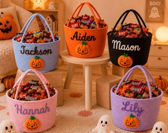 May include: Five colorful Halloween candy baskets filled with treats. Each basket is a different color: blue, orange, black, pink, and purple. Each basket has a name embroidered on it, along with a pumpkin applique.