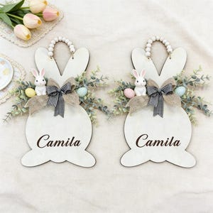 Puede incluir: Dos letreros de madera con forma de conejito de Pascua con el nombre "Camila" en escritura marrón. Cada letrero presenta una figura de conejito blanco, vegetación artificial, un lazo de cuadros y huevos decorativos. Un asa de cuentas remata cada letrero.