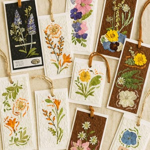 May include: A selection of handmade bookmarks, each adorned with pressed flowers. The bookmarks showcase diverse floral arrangements in pink, yellow, orange, blue, and white hues. Some have brown or black backgrounds, and all are finished with a hanging string.