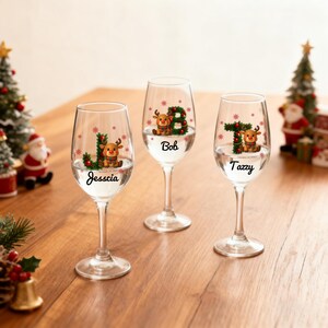 Puede incluir: Tres copas de vino transparentes con diseños navideños personalizados. Cada copa presenta una letra, un reno y un nombre: Jesscia, Bob y Tazz. Las copas están llenas de un líquido transparente y sobre una superficie de madera.