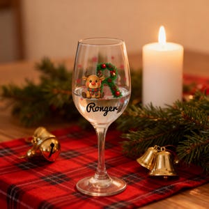 Könnte beinhalten: Ein klares Weinglas mit einem Weihnachtsrentier-Design und dem Namen "Ronger" aufgedruckt. Das Glas ist mit Flüssigkeit gefüllt und steht auf einer rot karierten Tischdecke, mit einer brennenden Kerze und Weihnachtsdekorationen im Hintergrund.