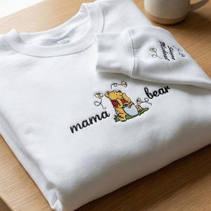 May include: White sweatshirt with the words "mama bear" embroidered in black script, along with a Winnie the Pooh graphic. The sleeve has the words "Summer Medical" embroidered in black script. A small plant and a mug are in the background.