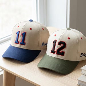 May include: Two baseball caps with contrasting brims and the numbers 11 and 12 embroidered on the front. The caps are beige with dark blue and green brims, respectively. The number 11 is in blue, and the number 12 is in red.