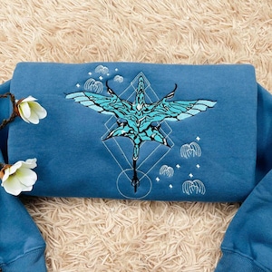May include: A blue sweatshirt with an embroidered design featuring a stylized bird with outstretched wings in shades of turquoise and white. The design is centered on the chest and includes geometric shapes and cloud-like elements. The sleeves are cuffed.