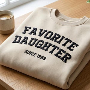 May include: A folded beige sweatshirt with the text "FAVORITE DAUGHTER" embroidered in black, with "SINCE 1999" below. The sweatshirt is on a wooden table, next to a mug of coffee and a small potted plant.