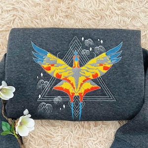 May include: Dark gray sweatshirt featuring a vibrant, embroidered design of a stylized bird with outstretched wings. The bird's wings are blue and yellow, with red and orange accents. The design is set against a white geometric shape.