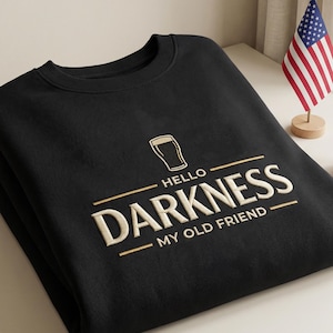 May include: Black sweatshirt with the text "HELLO DARKNESS MY OLD FRIEND" in a cream-colored, embroidered font. A small illustration of a beer glass is above the text. An American flag and a small potted succulent are in the background.