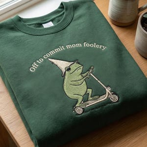 Könnte beinhalten: Dunkelgrünes Sweatshirt mit dem gestickten Schriftzug "Off to commit mom foolery". Das Design zeigt einen grünen Frosch mit spitzem Hut, der einen Roller fährt. Das Sweatshirt liegt auf einer Holzoberfläche.