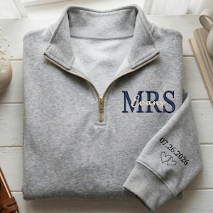 Puede incluir: Una sudadera gris con cremallera de un cuarto, cremallera dorada y texto bordado. El texto "MRS Evans" está en azul marino y dorado. La manga tiene la fecha "07.26.2026" y dos símbolos de corazón. La sudadera está doblada sobre una superficie blanca.