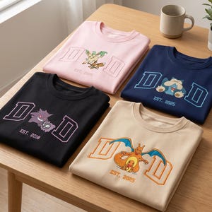Könnte beinhalten: Vier gefaltete T-Shirts in den Farben Rosa, Marineblau, Schwarz und Beige. Jedes Shirt zeigt das Wort "DAD" in einem Blockbuchstaben-Design, mit einer Cartoon-Figur-Grafik und dem Jahr "EST. 2025" oder "EST. 2018".