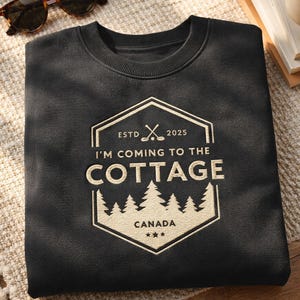 Może przedstawiać: Czarna bluza z kremową grafiką z napisem "I'M COMING TO THE COTTAGE" z kijem i piłką golfową, drzewami i słowem "CANADA". Bluza ma okrągły dekolt i długie rękawy. Okulary przeciwsłoneczne w lewym górnym rogu.