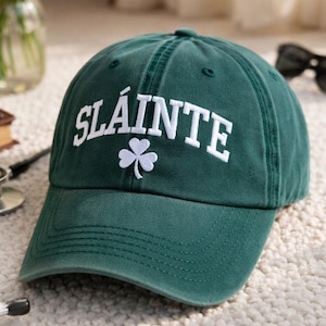 May include: A dark green baseball cap featuring the word "SLÁINTE" in white, with a white shamrock emblem. The cap has a curved peak and is made of a soft fabric. A casual hat.