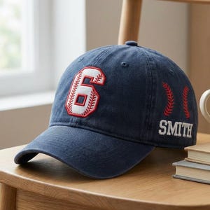 May include: Navy blue baseball cap with a white and red embroidered number 6 on the front. The name "SMITH" is embroidered in white on the side, with red baseball stitching details. The cap is sitting on a wooden chair.