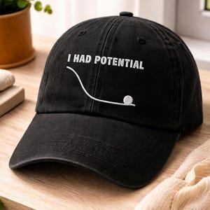 Pode incluir: Boné de beisebol preto com a frase bordada "I HAD POTENTIAL" acima de um gráfico branco de uma bola de golfe num tee. O boné é feito de um tecido macio e tem uma aba curva.