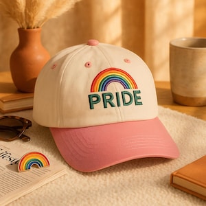 Può includere: Un cappellino da baseball con corona color crema e visiera rosa. Il cappellino presenta un arcobaleno ricamato e la parola "PRIDE" in verde. È visibile anche una piccola spilla arcobaleno.