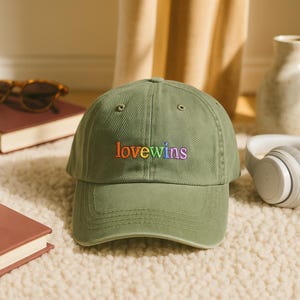 May include: Olive green baseball cap with the embroidered text "lovewins" in rainbow colors. The cap has a curved brim and a classic six-panel design. The hat is sitting on a fluffy, off-white rug.