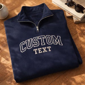 May include: Navy blue fleece pullover with a quarter-zip collar. The embroidered text "CUSTOM TEXT" is in white block letters. Sunglasses and books are in the background.