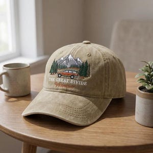 May include: A tan baseball cap with an embroidered design featuring a mountain scene, trees, and a vintage station wagon. The text "THE GREAT DIVIDE VERMONT" is arched below the image. The cap is sitting on a wooden table.