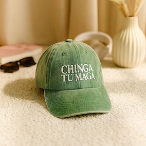 Può includere: Cappellino da baseball verde con la scritta "CHINGA TU MAGA" in bianco. Il cappellino ha una visiera curva e un bottone sulla parte superiore. Il cappello è su una superficie strutturata.
