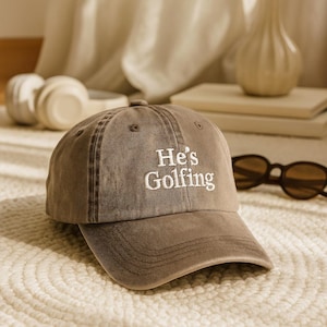 May include: A brown baseball cap with the embroidered text "He's Golfing" in white. The cap has a curved brim and a classic six-panel design. Sunglasses are visible in the background.