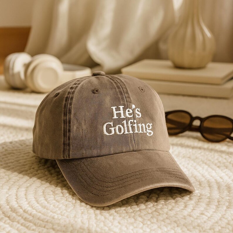 May include: A brown baseball cap with the embroidered text "He's Golfing" in white. The cap has a curved brim and a classic six-panel design. Sunglasses are visible in the background.