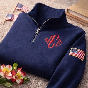 May include: Navy blue quarter-zip sweatshirt with a red embroidered monogram and an American flag patch on the sleeve. The sweatshirt has a high collar and a silver zipper. Two small American flags and flowers are in the background.