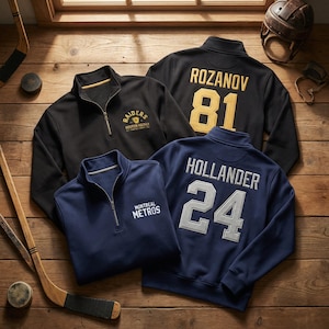 May include: Four quarter-zip pullovers in black and navy blue, with hockey-themed logos and player names and numbers. The black pullovers feature "Raiders" and "Rozanov 81". The navy blue pullovers display "Montreal Metros" and "Hollander 24". Hockey sticks and a helmet are in the background.