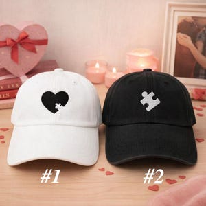 May include: Two baseball caps, one white and one black. The white cap features a black heart with a puzzle piece, and the black cap has a white puzzle piece. The numbers "#1" and "#2" are printed below the caps.