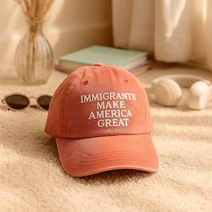 Puede incluir: Gorra de béisbol color coral con el texto blanco "IMMIGRANTS MAKE AMERICA GREAT". La gorra está sobre una alfombra crema, con gafas de sol y auriculares en el fondo. Un accesorio casual.