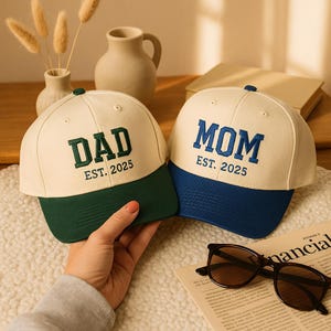 Może przedstawiać: Dwie czapki bejsbolowe, jedna kremowo-zielona, druga kremowo-niebieska. Na pierwszej czapce napis "DAD" wyszyty na zielono z napisem "EST. 2025" poniżej. Druga czapka ma napis "MOM" w kolorze niebieskim z napisem "EST. 2025".