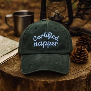May include: A dark green baseball cap with the embroidered text "Certified Napper" in light blue. The hat is on a wooden surface, with a metal mug, map, lantern, and pine cones in the background.