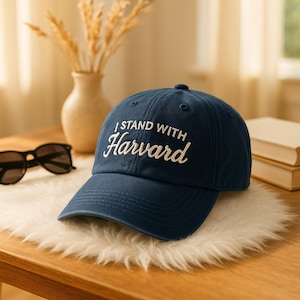 May include: A navy blue baseball cap with the embroidered text "I STAND WITH Harvard" in white script. The cap is sitting on a white, fluffy surface, with sunglasses and a vase in the background. The hat has a curved brim.