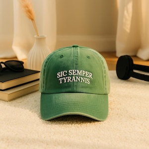 May include: Green baseball cap with the text "SIC SEMPER TYRANNIS" in white. The cap is made of a soft material and has a curved brim. The cap is sitting on a white, textured surface.