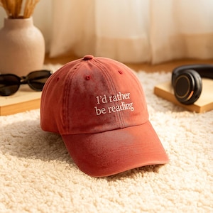 May include: A red baseball cap with the text "I'd rather be reading" in white. The cap is made of a soft fabric and has a curved brim. The cap is sitting on a white, fluffy rug.