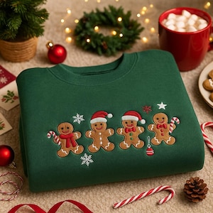 Może przedstawiać: Zielona bluza z haftowanym wzorem piernikowych ludzików w czapkach Świętego Mikołaja i trzymających laski cukrowe. Projekt zawiera płatki śniegu i gwiazdę. Świąteczny motyw.