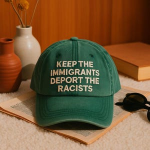 Puede incluir: Gorra de béisbol verde con el texto "KEEP THE IMMIGRANTS DEPORT THE RACISTS" en blanco. La gorra está sobre una superficie estampada, con gafas de sol y un libro al fondo. La gorra tiene visera curva.