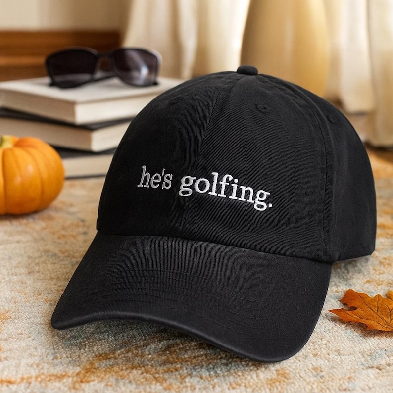 May include: Black baseball cap with the text "he's golfing" embroidered in white. The cap has a curved brim and a classic six-panel design. The hat is sitting on a patterned surface.