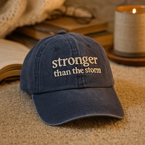 Op de afbeelding: Een marineblauwe baseballcap met de geborduurde tekst "stronger than the storm" in het wit. De cap ligt op een beige tapijt, met een brandende kaars en boeken op de achtergrond.