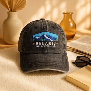May include: A dark gray baseball cap with a mountain and star design. The cap features the text "VELARIS CITY OF STARLIGHT" embroidered on the front. The design includes blue mountains, green trees, and yellow stars. The cap has a curved brim and a button on top.