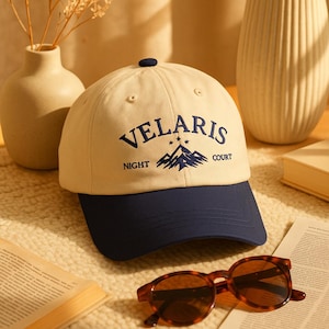 May include: A beige baseball cap with a navy blue brim and top button. The cap features the text "VELARIS NIGHT COURT" above a mountain graphic in navy blue. Sunglasses and a book are in the foreground.