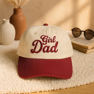 Puede incluir: Gorra de béisbol crema con visera burdeos. La gorra presenta las palabras bordadas "Girl Dad" en una fuente cursiva. La gorra está sobre una superficie blanca y esponjosa.