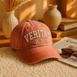 May include: A dusty rose baseball cap with the word "VERITAS" embroidered in off-white, along with the number "1636". The cap is sitting on a cream-colored, textured surface.