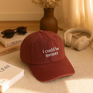 Könnte beinhalten: Burgunderfarbene Baseballkappe mit dem weißen Schriftzug "i could be meaner". Auf einer cremefarbenen Oberfläche liegen Sonnenbrille, ein Buch, eine Vase und Kopfhörer. Das Ensemble befindet sich auf einem Teppich.